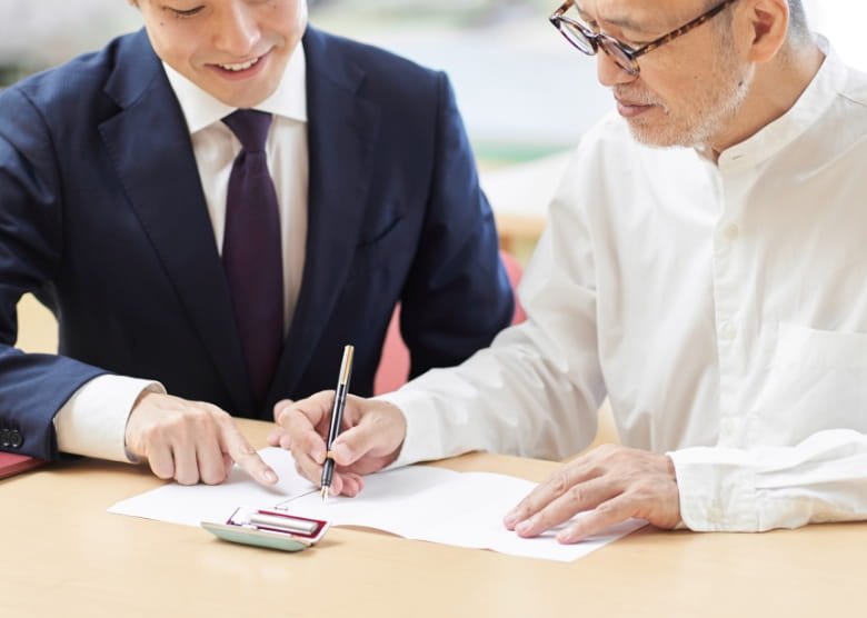スーツ姿の男性が高齢の男性に書類への署名を案内している様子